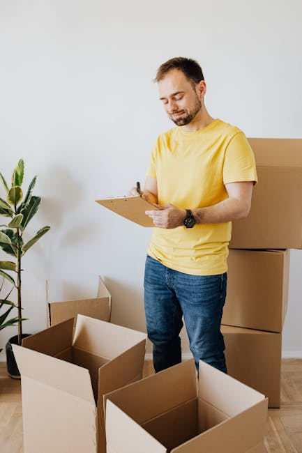 A man with short dark hair and a beard, dressed in a yellow t-shirt and blue jeans, is standing indoors in front of a plain white wall. He is holding a clipboard and pen, appearing to take notes or pe