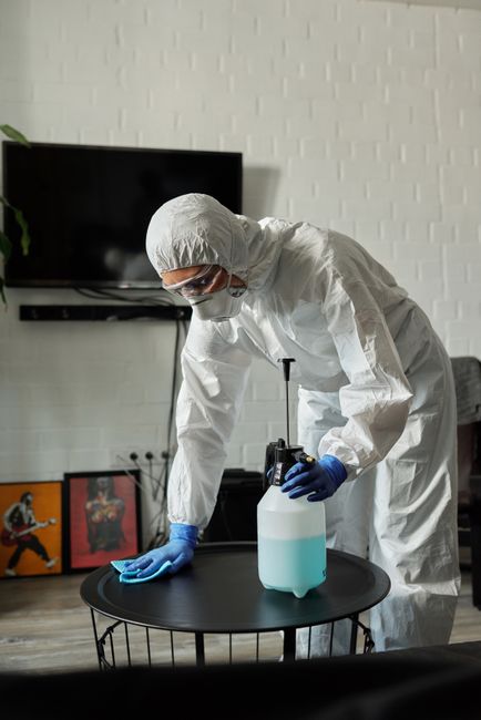 A professional cleaner dressed in full white protective coveralls, face mask, goggles, and blue gloves is performing surface cleaning in a modern indoor setting. They are using a spray bottle filled w