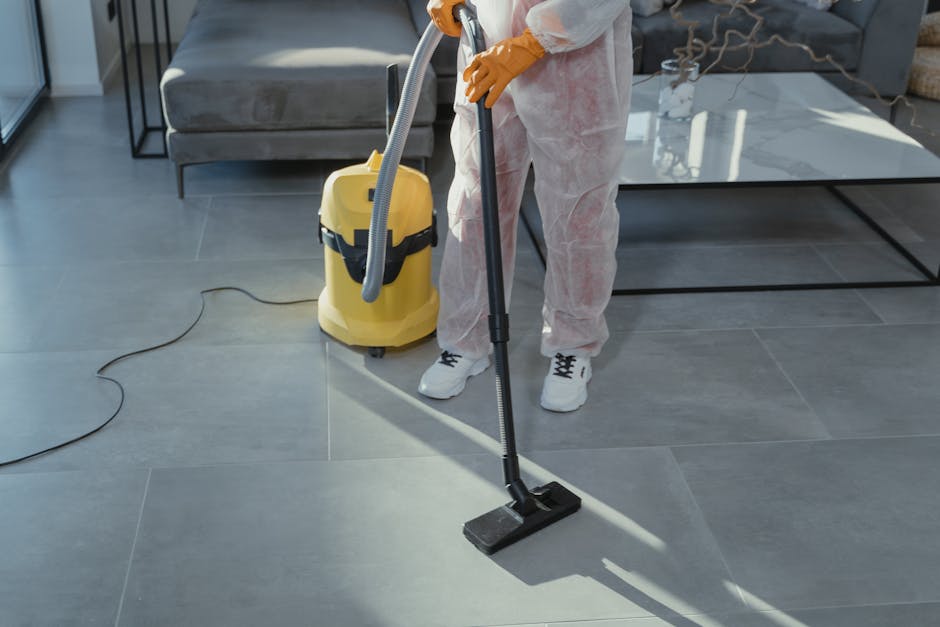 A professional cleaner dressed in full white protective coveralls, face mask, goggles, and blue gloves is performing surface cleaning in a modern indoor setting. They are using a spray bottle filled w